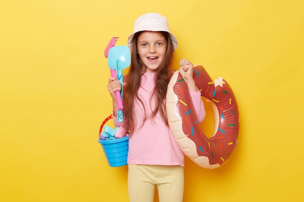 Extremely happy overjoyed little girl in panama holding sandbox toys bucket and rubber ring isolated over yellow background rejoicing coming to beach