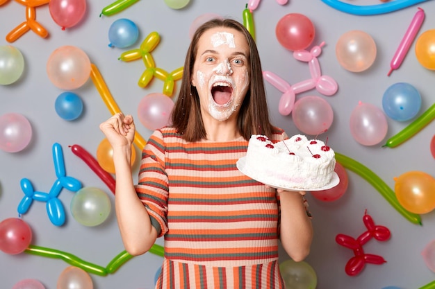 Extremely happy overjoyed woman in striped dress standing against gray wall decorated with colorful balloons holding cake being dirty in cream clenched fist screaming