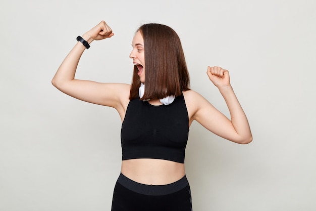 Extremely happy slim woman with headphones wearing sportswear posing against gray background looking at her biceps with amazed excited expression