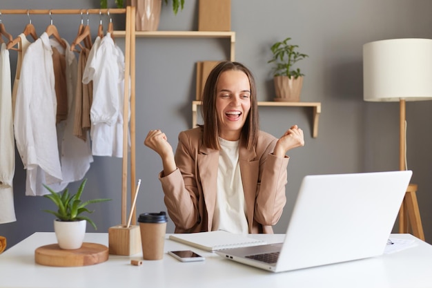 Extremely happy woman designer or stylist in jacket working on laptop computer while sitting on table against clothes hang on shelf clenched fists celebrating her success finishing new collection