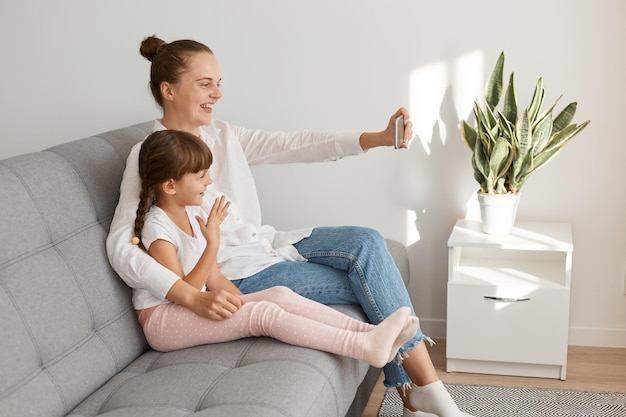 Extremely happy woman posing in living room with her daughter and having video call or taking selfie, looking at cell phone camera, family wearing casual style clothing.