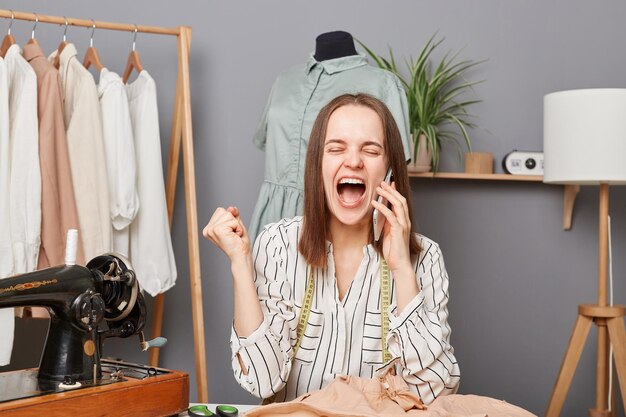 Extremely happy woman seamstress smiling talking on mobile phone in her sewing workshop clenched fists rejoicing her success screaming with happiness finishing new collection