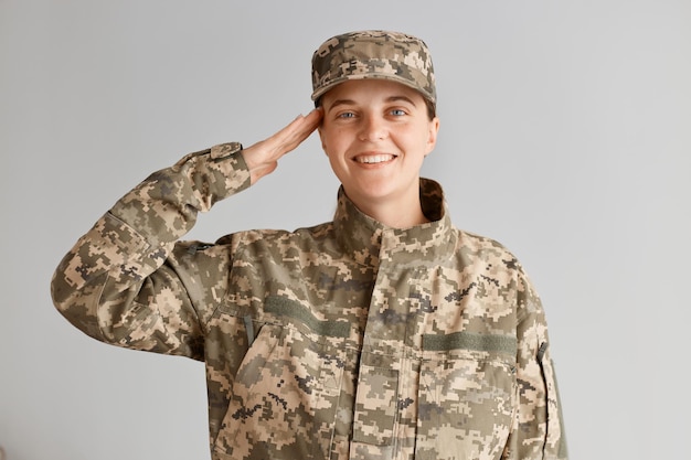 Extremely happy woman soldier with toothy smile and optimistic emotions saluting while standing against light background indoor, wearing camouflage uniform and cap.