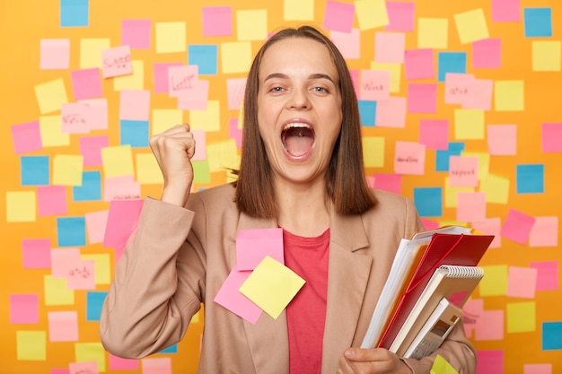 Photo extremely happy woman wearing beige jacket standing with paper folders clenched fists celebrating to win tender screaming with happiness posing over sticky notes to write reminder on yellow wall