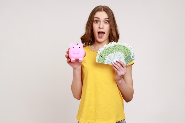 Чрезвычайно потрясенная женщина молодого возраста в желтой футболке с копилкой и банкнотами евро, смотрящая в камеру с открытым ртом, депозитный кэшбэк. Снимок в помещении, изолированный на сером фоне