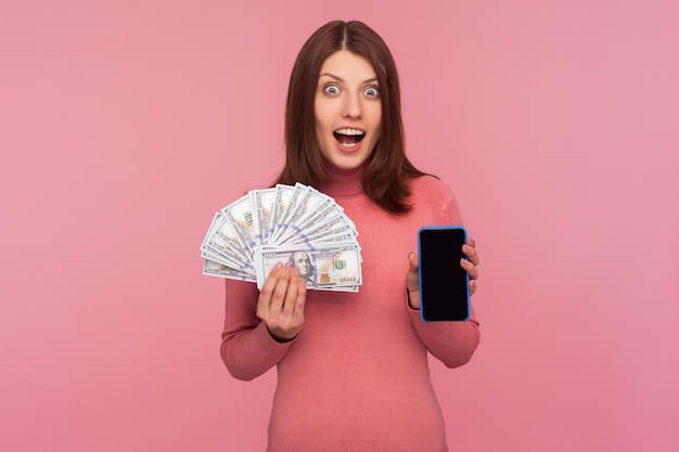 Extremely surprised excited brunette woman in pink sweater holding smartphone with empty screen and dollar bills looking at camera with shock Indoor studio shot isolated on pink background