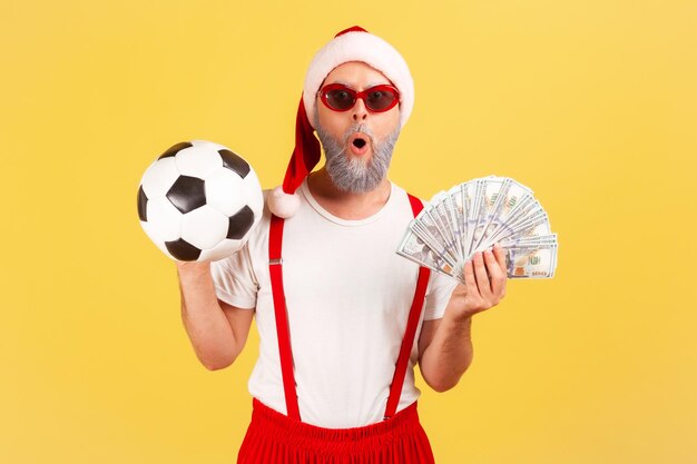 Фото Чрезвычайно удивленный потрясенный пожилой мужчина в шляпе санта-клауса и стильных очках, держащий долларовые банкноты и футбольный мяч, делающий ставки на спорт, победу. крытая студия снята на желтом фоне