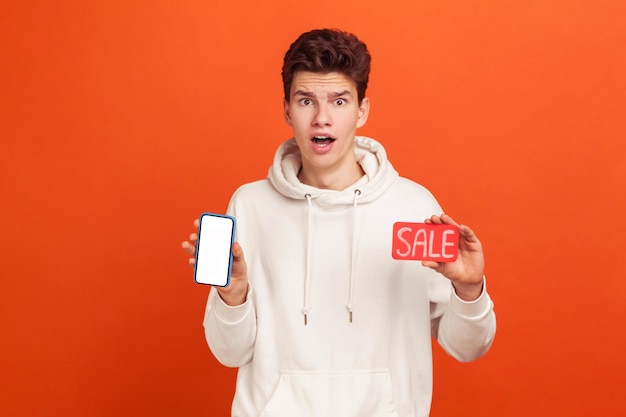 Extremely surprised teenager in casual hoodie widely open eyes and mouth holding sale card and smartphone  shocked with amazing online sellout Indoor studio shot isolated on orange background