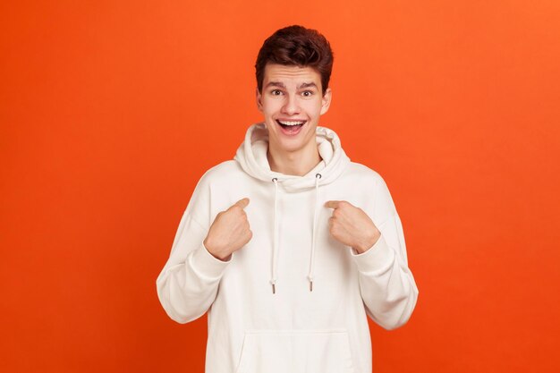 Extremely surprised teenager in white casual style hoodie pointing fingers on himself with widely opened mouth and eyes shocked with victory unbelievable Indoor isolated on orange background