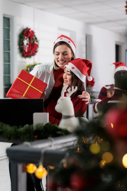 Exuberant worker receiving gift from colleague during Christmas holiday season. Excited employee gets red box present from coworker on secret santa party in festive decorated workspace