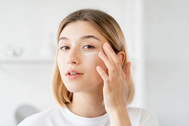 Under eye cream Skincare moisturizing Morning treatment Pretty young woman applying beauty product on soft face skin at light bathroom