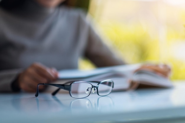 An eye glasses on the table with blurred of a woman reading book in background