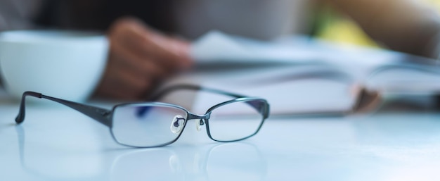 An eye glasses on the table with blurred of a woman reading book in background