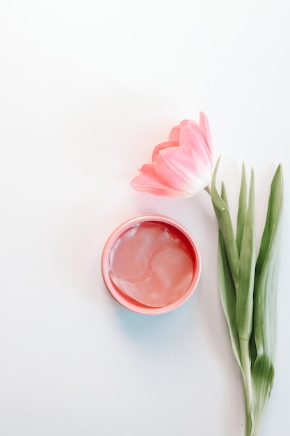 Under eye patches in jar and flowers on white background flat lay Space for text