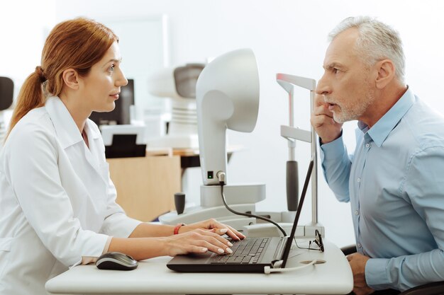 Eye specialist smiling. Pleasant eye specialist smiling while examining aged grey-haired man