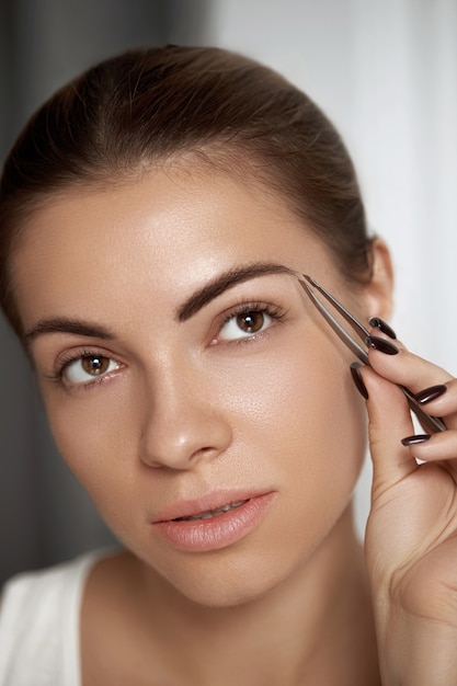 Eyebrow correction. plucking eyebrows. beautiful young woman\
with tweezers. model with beauty face.