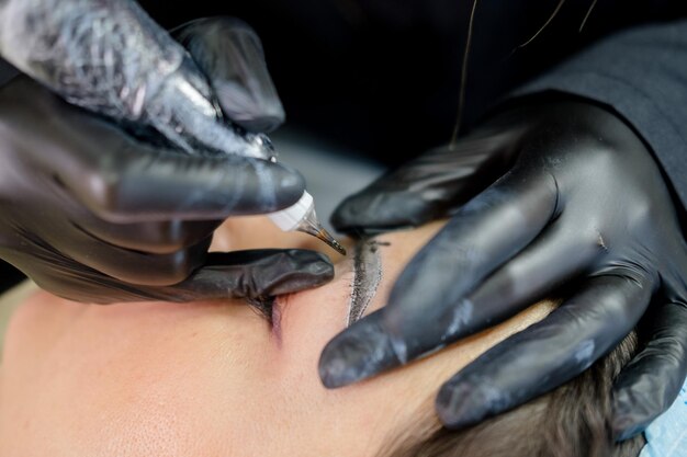 Eyebrow microblading. A master in black gloves holds a blending needle over the brow of the model.