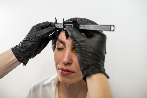 Photo eyebrow reshaping stylist measuring eyebrows with a ruler cosmetician makes measuring before permanent brow makeup to a glorious brunette woman in cosmetology salon