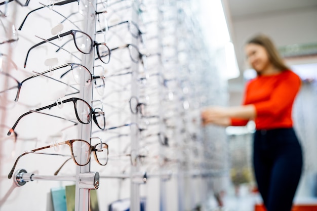 Eyeglasses shop. Stand with glasses in the store of optics. Showcase with spectacles in modern ophthalmic store. Closeup. Woman on blurred background.