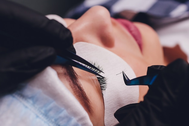 Eyelash Extension Procedure. Close up view of female eye with long eyelashes. Stylist holding pink tweezers, tongs and making lengthening lashes. Macro, selective focus. Beauty Concept. Treatment.