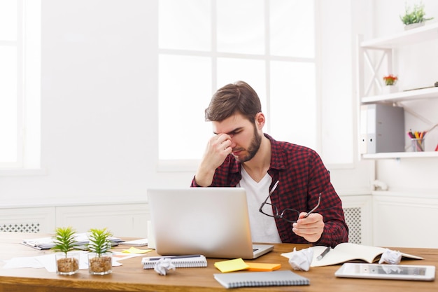 Eyes pain. Tired, overworking businessman with laptop in modern white office interior. Handsome man in casual at work with computer, job problems concept.