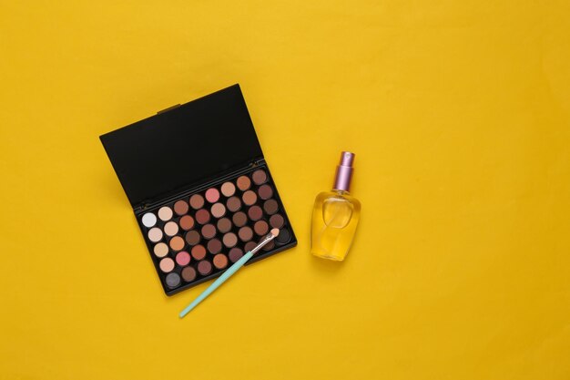 Eyeshadow makeup palette and perfume bottle on yellow background Beauty and fashion scene Top view