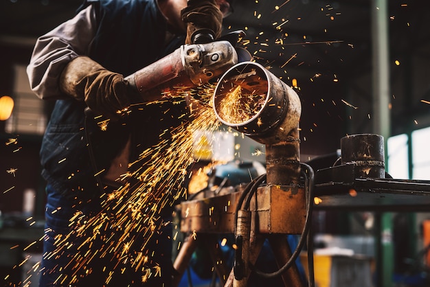 Fabric worker in protective uniform cutting metal pipe on the work table with an electric grinder in the industrial workshop.