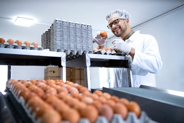 Fabrieksarbeider in witte jas en haarnetje die de kwaliteit controleert van industrieel geproduceerde eieren in een voedselverwerkende fabriek of boerderij.