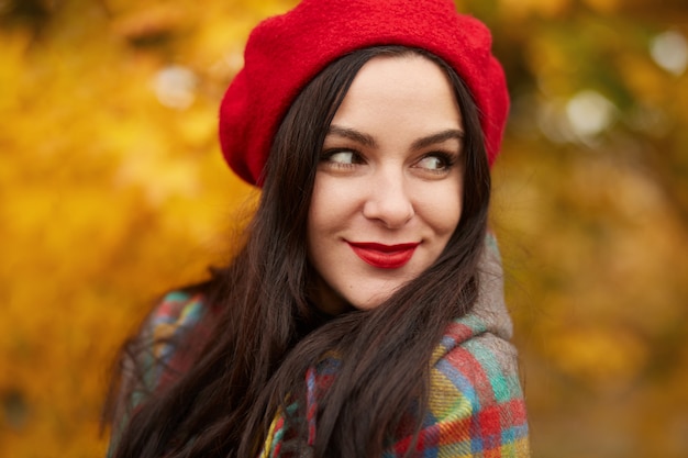 Fabulous romantic woman with long dark hair wearing red beret on blurred autumn forest. Girl in forest with orange autumn leaves