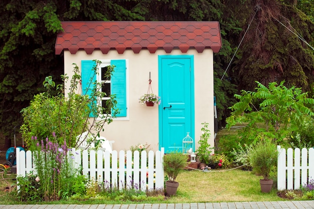 Facciata di piccola casa con finestra e porta aqua, fiori in vaso sul muro e vista sul giardino