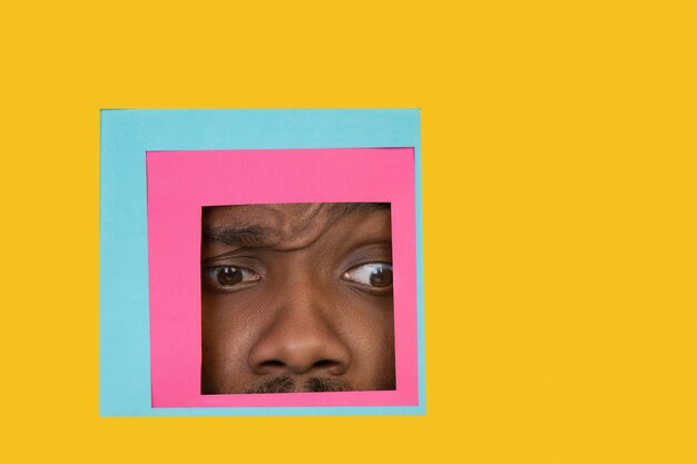 Face of african-american man peeking throught square in yellow background