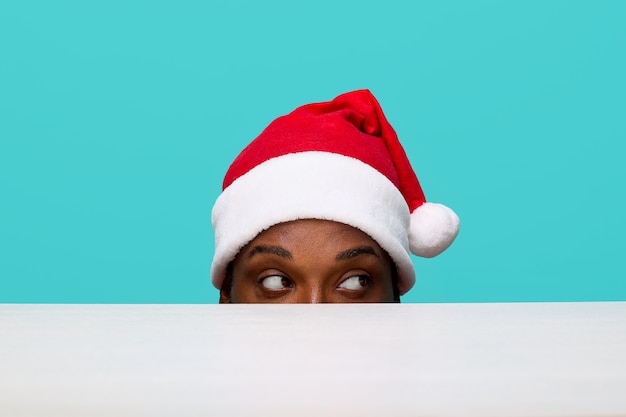 Face of an AfricanAmerican guy in a Santa Claus hat looking from under the table to the right