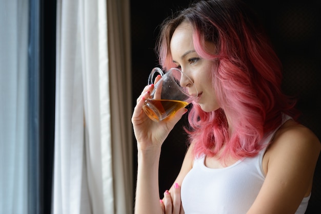 Лицо красивой женщины с розовыми волосами, пьющей чай и смотрящей в окно