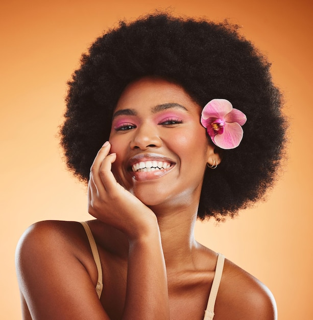 Photo face beauty and black woman with flower in afro hair on orange studio background portrait skincare and happy woman or female model from nigeria in pink makeup cosmetics and orchid in healthy hair