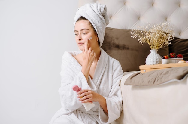 Face cream woman in bathrobe with a towel on her head after showering at home. applying skin cream on skin.