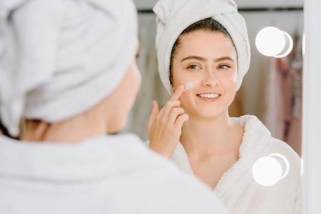 Face cream woman in bathrobe with a towel on her head after showering at home. applying skin cream on skin.