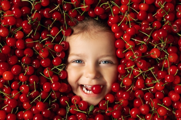 The face of a happy cheerful child surrounded by a large number of red ripe juicy cherries