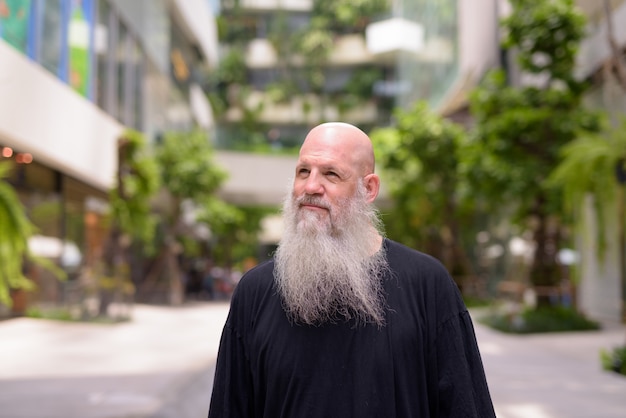 Face of happy mature handsome bald bearded man thinking with nature in the city