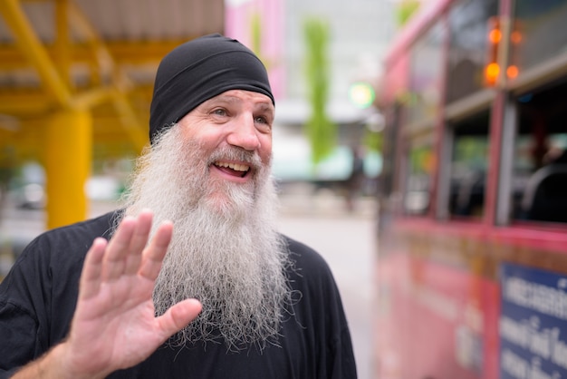 Face of happy mature handsome bearded hipster man waving hand at the bus