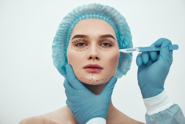 化粧品の顔を持っている青い医療帽子の美しい若い女性のフェイスリフト手術の肖像画