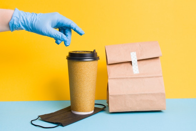 face mask, coffee and package with food for delivery, hand in glove on yellow background contactless delivery concept