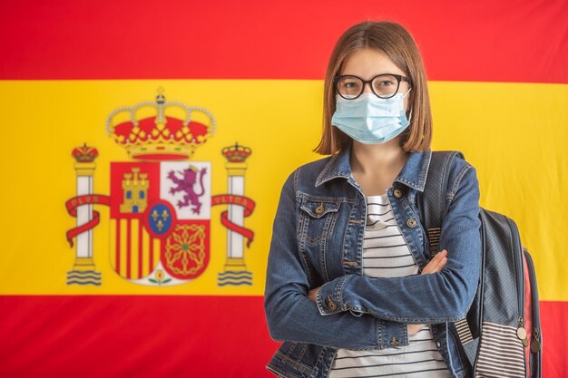 Face mask wearing female student carrying backpack with spanish flag on the background