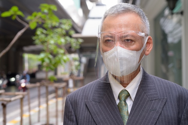 Face of mature Japanese businessman with mask and face shield thinking in the city