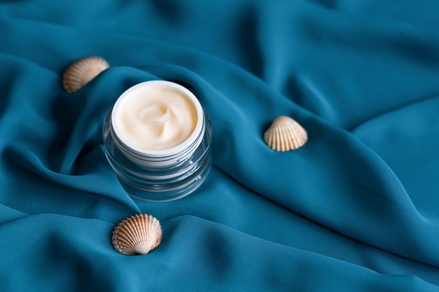 Crema idratante minerale viso con conchiglie in vasetto di vetro su seta blu.