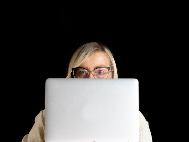 Фото Лицо зрелой женщины с очками выглядит из-под ноутбука, изолированного на черном фоне