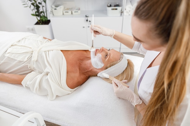 Face peeling mask, spa beauty treatment, skincare. Senior woman getting facial care by beautician at spa salon.