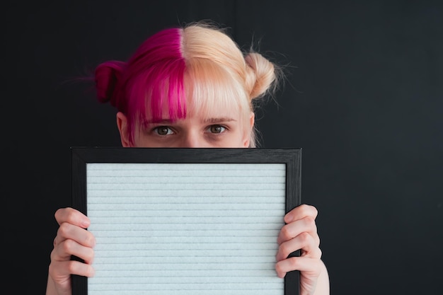 face of pink haired girl holding empty frame in hands on black background