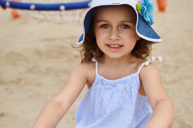 Face portrait of a cute beautiful little girl wearing sunhat and relaxing on the sand. Summer camp