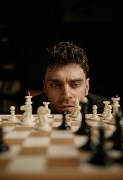 Face portrait of thoughtful young man player considering chess piece position on board planning strategic move. Selective focus and closeup view