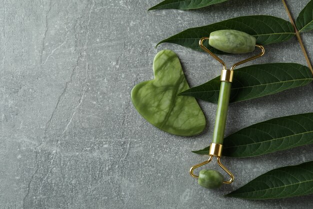 Face roller, gua sha and twig on gray textured background